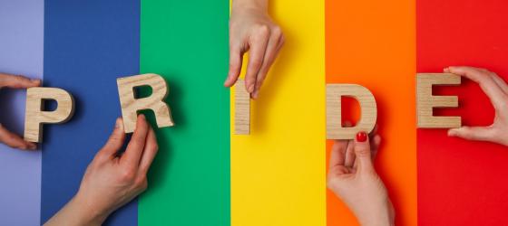 scritta PRIDE in singole lettere di legno appoggiate da alcune mani su uno sfondo arcobaleno