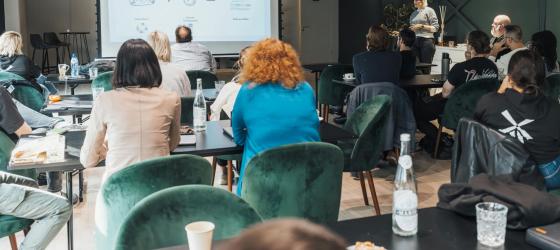 Aula moderna con persone sedute a tavoli neri e sedie verde scuro, mentre assistono a una presentazione proiettata su uno schermo. Una donna in piedi, vicino allo schermo, sta parlando al gruppo. L’atmosfera è informale e collaborativa, con bottiglie d’acqua, tazze e quaderni sui tavoli.