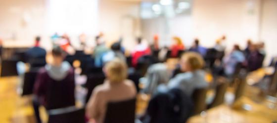 immagine sfuocata di una conferenza, molte persone sedute e sullo sfondo una o due persone che parlano