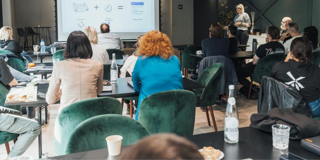 Aula moderna con persone sedute a tavoli neri e sedie verde scuro, mentre assistono a una presentazione proiettata su uno schermo. Una donna in piedi, vicino allo schermo, sta parlando al gruppo. L’atmosfera è informale e collaborativa, con bottiglie d’acqua, tazze e quaderni sui tavoli.